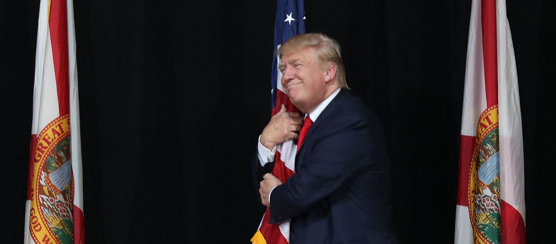 Тампа, Флорида - 24 октября: Кандидат в президенты от республиканцев Дональд Трамп обнимает американский флаг, когда он прибывает на митинг кампании в амфитеатре кредитного союза MidFlorida 24 октября 2016 года в Тампе, Флорида. До президентских выборов осталось 14 дней. Джо Рэдл / Getty Images / AFP (Фото Джо Рэдла - Sputnik Абхазия, 1920, 03.04.2021
