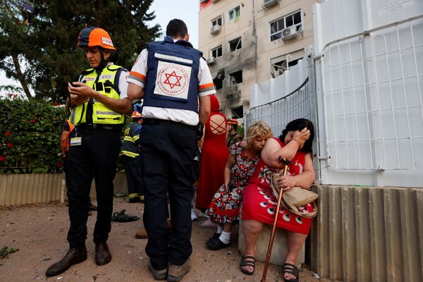 Израильские медики стоят рядом с женщинами возле дома, поврежденного ракетой, запущенной из сектора Газа в Ашкелоне - Sputnik Абхазия