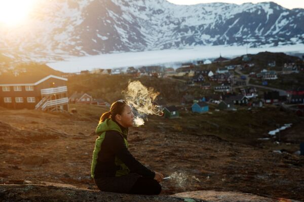 Молодая девушка сидит на камне и курит в лучах закатного солнца в городе Тасиилак, Гренландия - Sputnik Абхазия